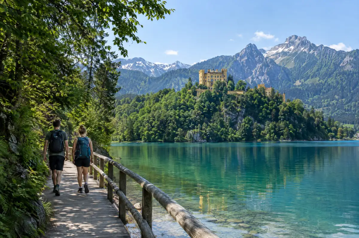 Two travelers walking the Alpsee lakeside path with Hohenschwangau Castle and the Alps behind