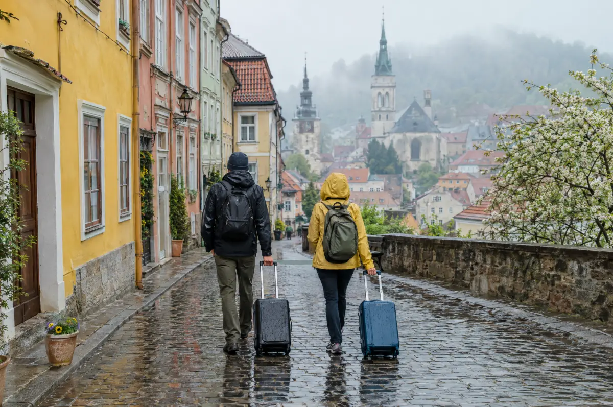 Two travelers in rain jackets pulling carry-on bags along a wet cobblestone street in an old European town